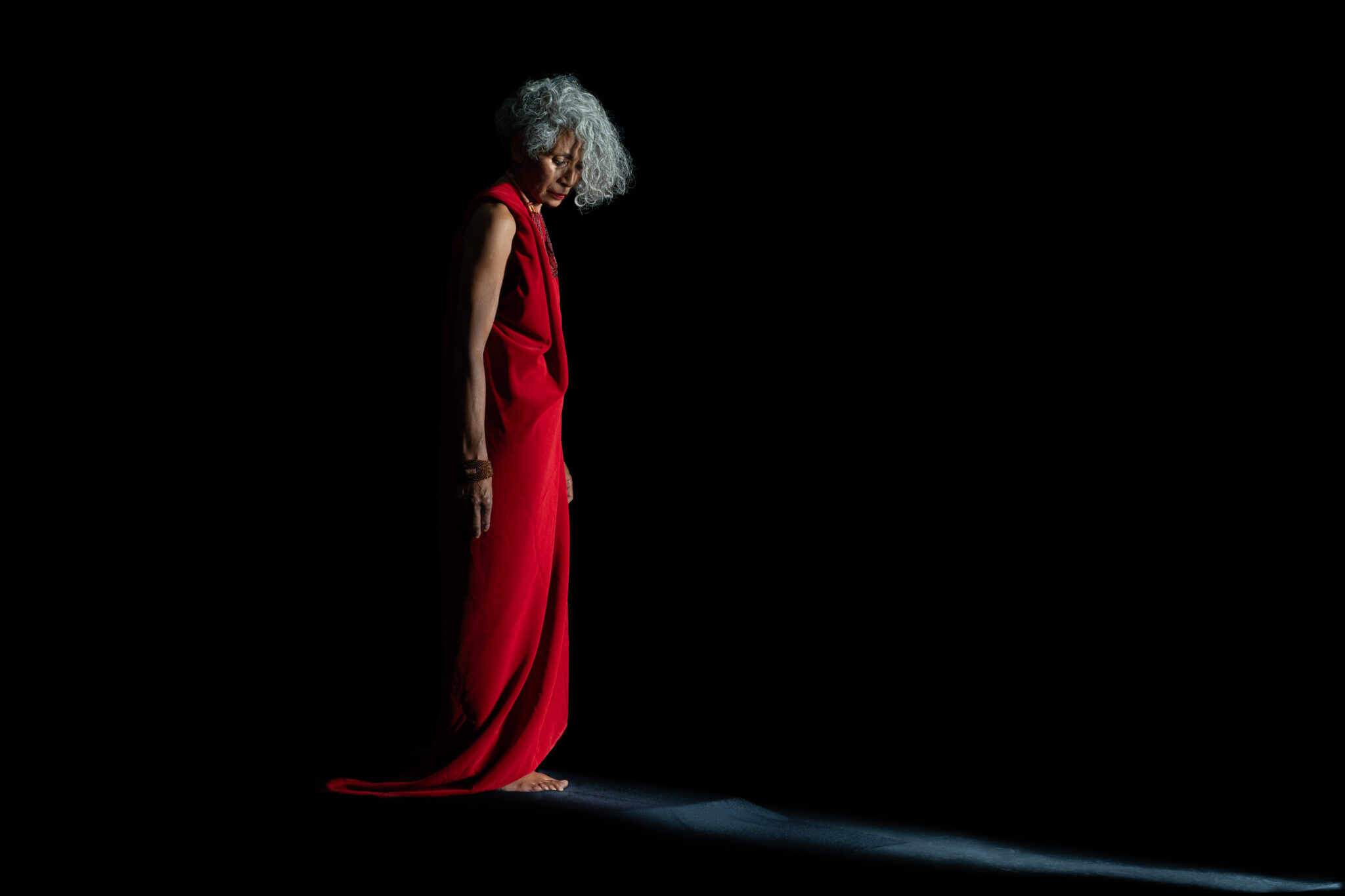 Woman standing in a red draped dress against a black background, head lowered, with a narrow band of light across the floor.