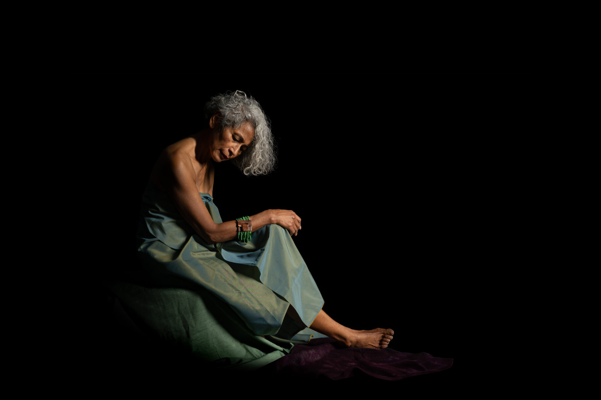 Woman seated and leaning forward in a pale green draped dress against a black background, head lowered and hands resting on one knee.