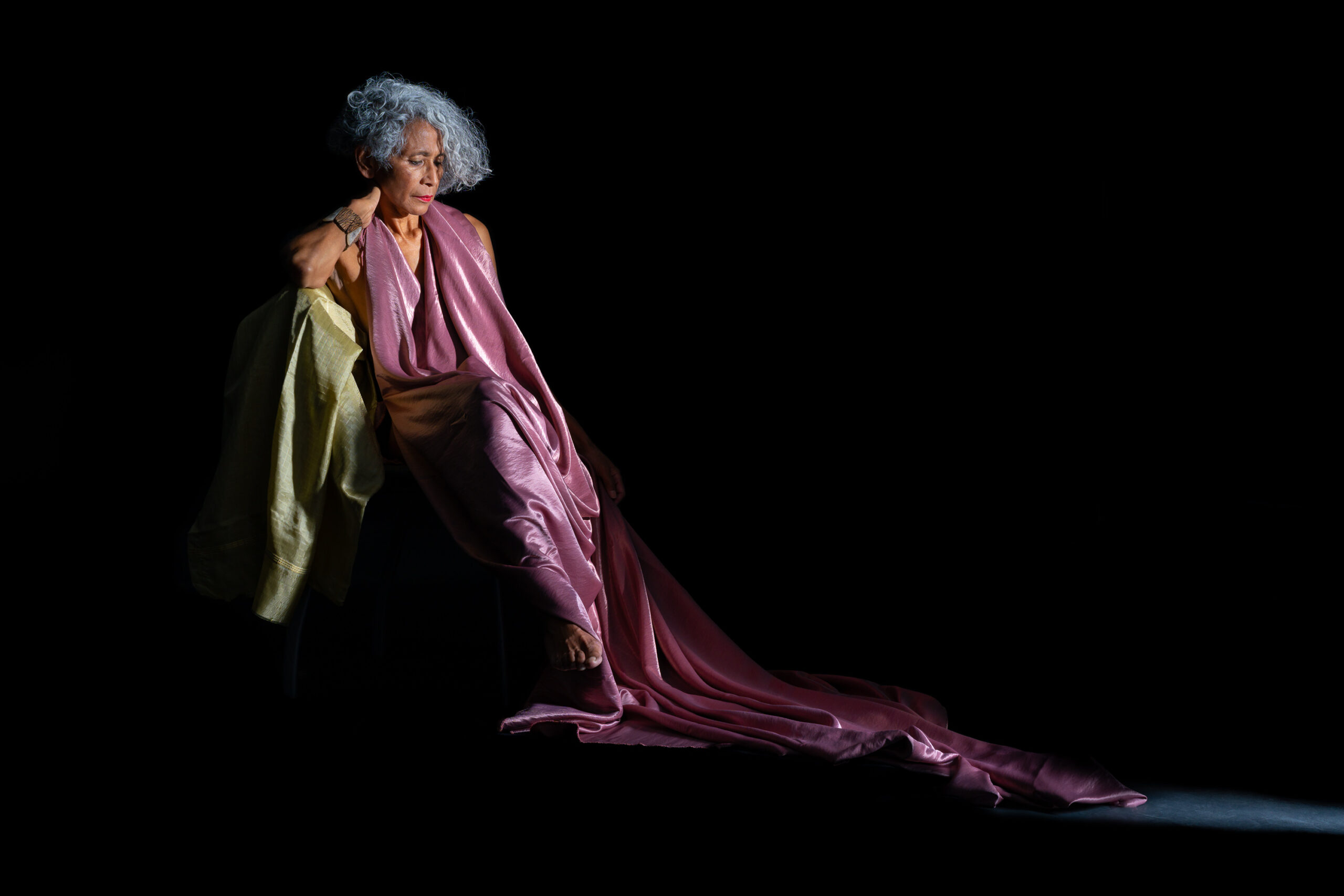 Woman seated in a purple draped garment against a black background, with one hand at her shoulder and her gaze lowered.