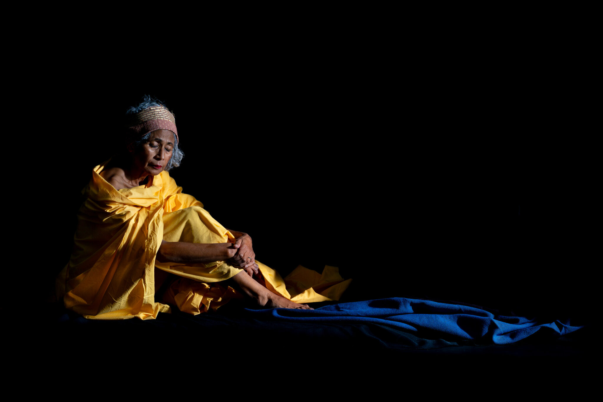 Woman seated in yellow fabric against a black background, wearing a head covering, with blue fabric extending across the foreground.