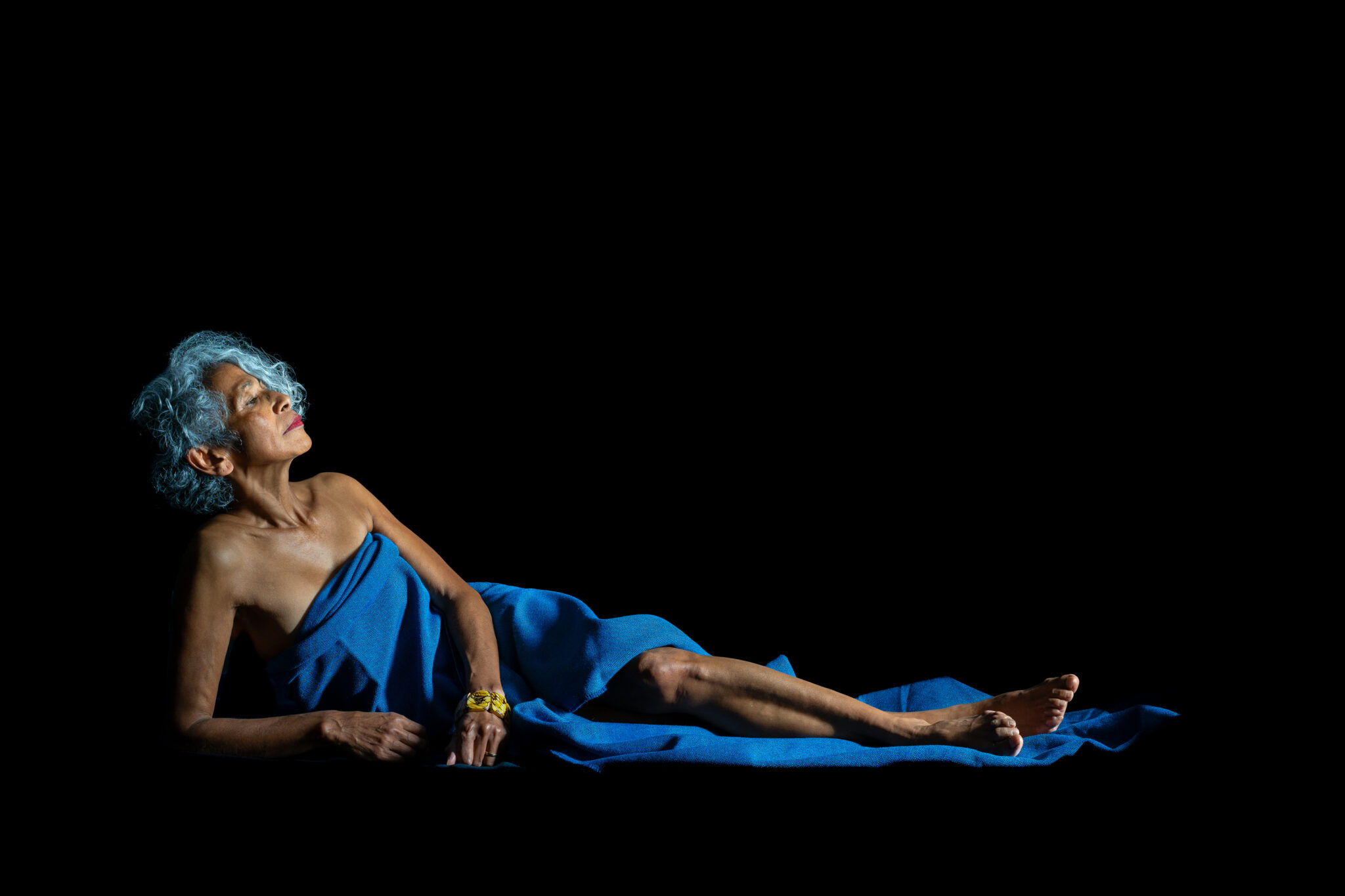 Woman reclining in blue fabric against a black background, head tilted upward into the light and one leg extended.